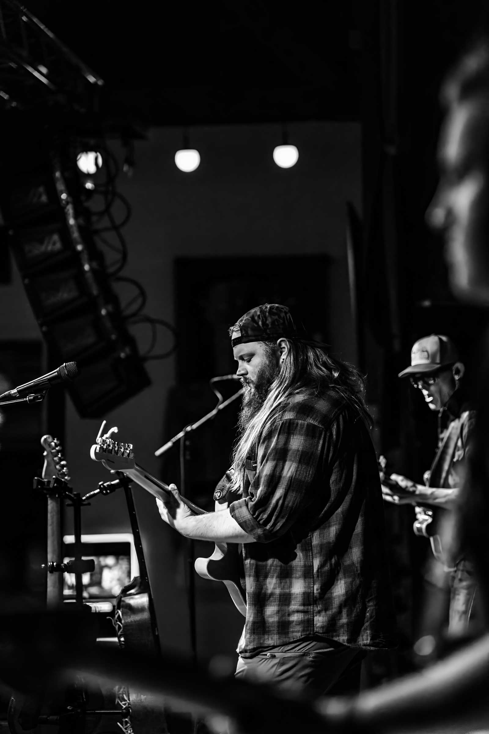 A black-and-white photo of a bearded man wearing a backwards cap and plaid shirt, playing an electric guitar on stage. Another musician, also playing a guitar, is visible in the background, both illuminated by stage lights.