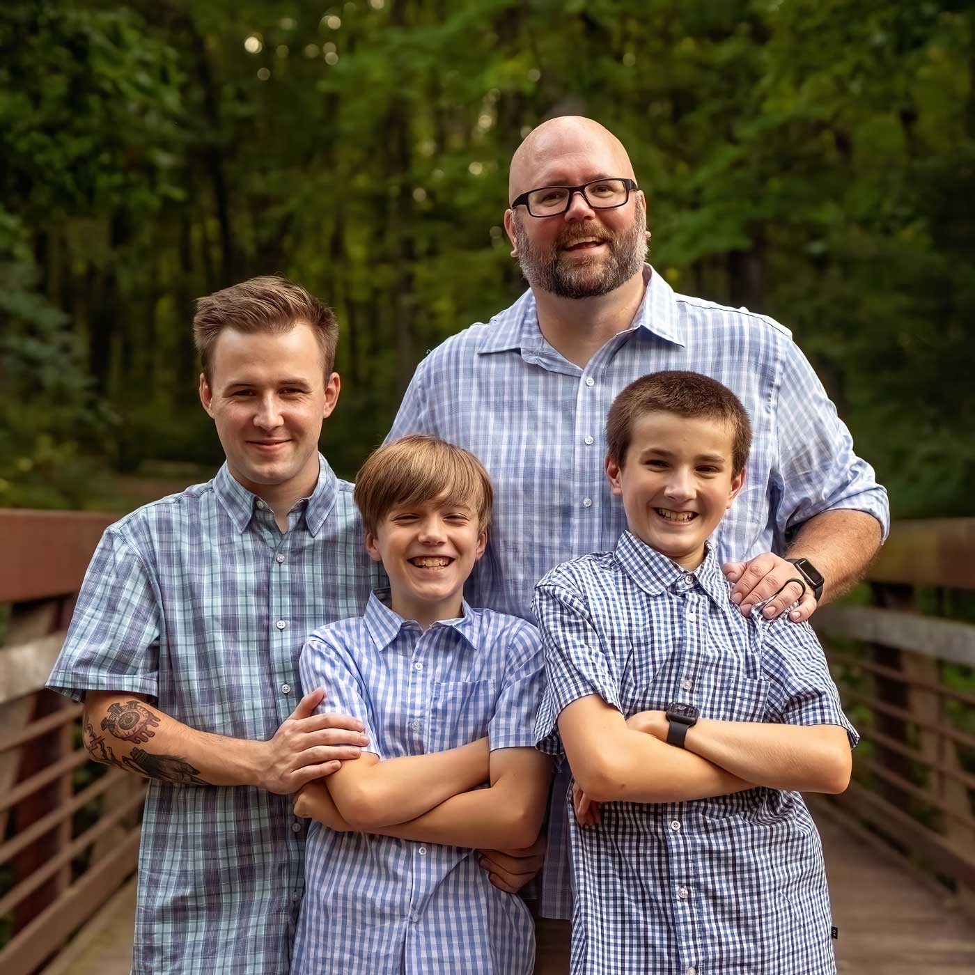 A family portrait taken outdoors on a bridge, featuring a bald man with glasses and a beard, standing behind three boys. All are wearing plaid shirts and smiling, with a wooded area in the background.