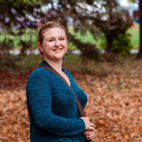 A photo of Summer Gladwell smiling towards the camera while standing in a wooded area with a strap over her shoulder.